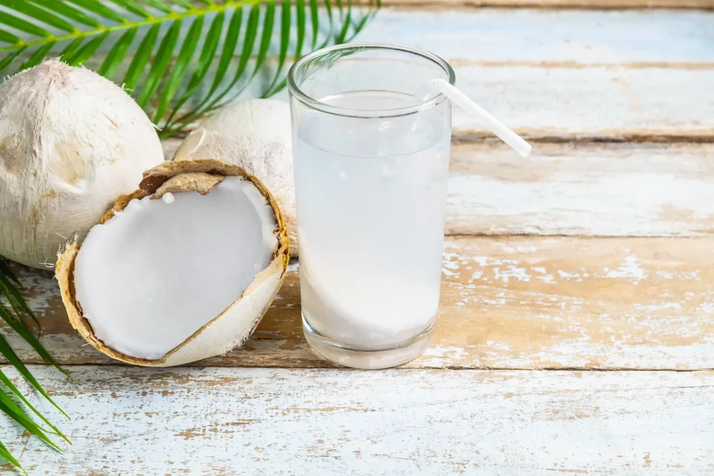 A tall, clear glass of coconut water with a straw in it sits on a table next to an open coconut. Coconut water and other summer drinks like these are good for after a spa soak.

