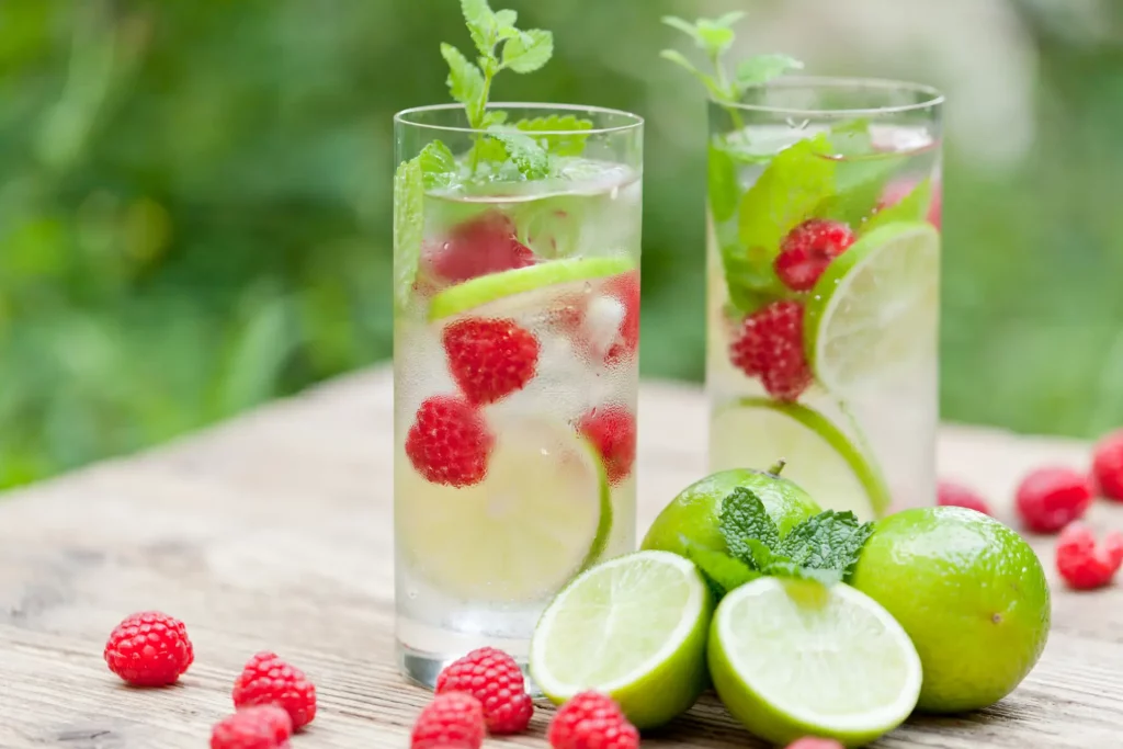 Two tall, clear glasses on a table contain water infused with lime, raspberries, and fresh herbs. Infused water and other summer drinks like these are good for after your spa soak.