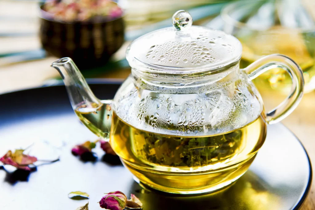 A clear, glass tea pot containing loose leaf tea steeping in hot water sits on a tray. Noncaffeinated herbal tea and other summer drinks like these are good for after a spa soak.
