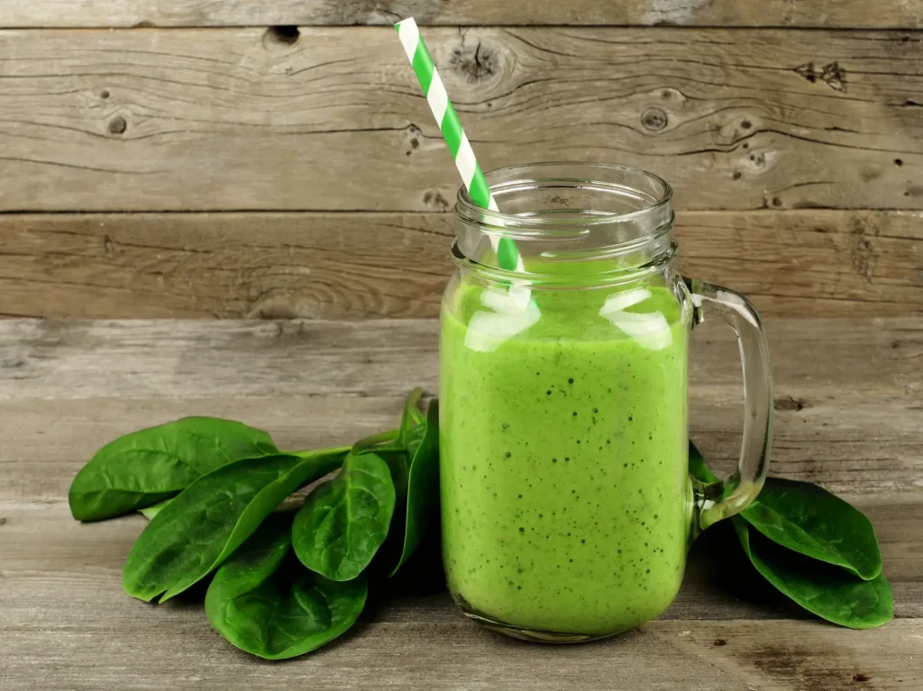 A green smoothie in a mason jar with a straw in it sits on a table next to a few leaves of baby spinach. Smoothies and other summer drinks like these are good for after a spa soak.