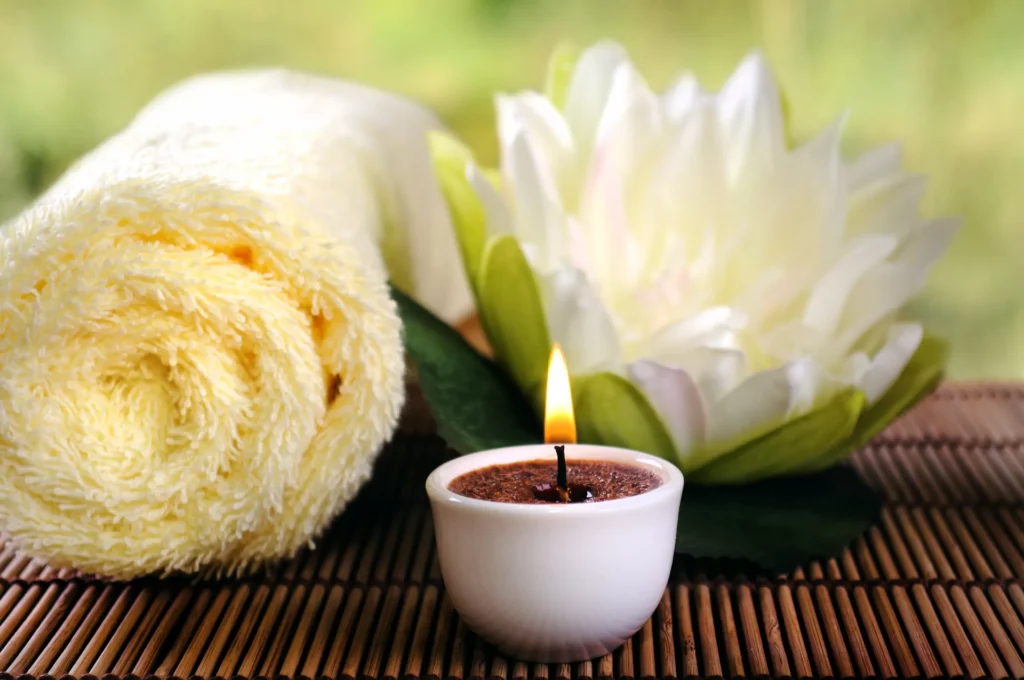 Close up of a rolled up, yellow towel, a white lily, and a small aromatherapy candle rest on a bamboo mat. Gathering products like these before your hot tub soak are one way to enhance the experience.