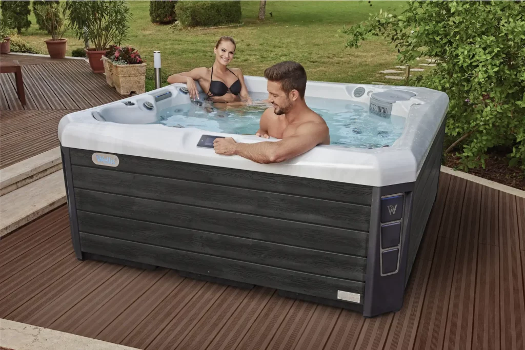 Two swimmers sit in a Wellis hot tub on a deck outdoors. Keeping water flowing through a tub like this is one of the key topics addressed through the question "Is hot tub maintenance hard?"
