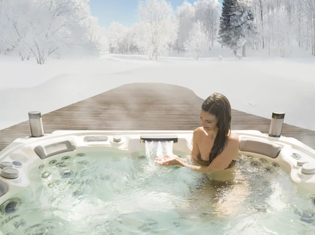 A swimmer sits in a Wellis hot tub in the midst of a snowy landscape. Using hot tubs and swim spas like these in winter requires knowledge of how to remove snow from a spa cover.
