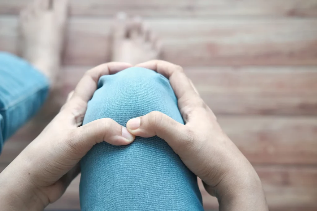 Close up of a person's knee, clothed by jeans, as the person rubs the joint. Knee stiffness and swelling are two symptoms that prompt the question of How Using a Sauna Provides Joint Pain Relief.