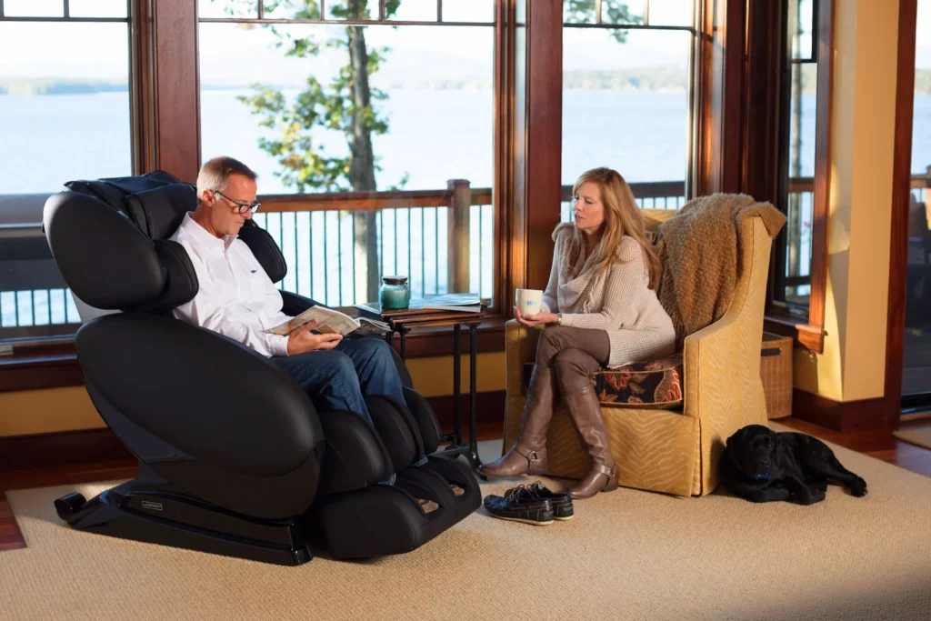 Medium shot of a couple seated in their home. One person sits in an Infinity Massage Chair while the other sits in a cushioned chair. Infinity Massage Chairs are one of the wellness solutions you can bring home during the holidays to improve your overall well-being.