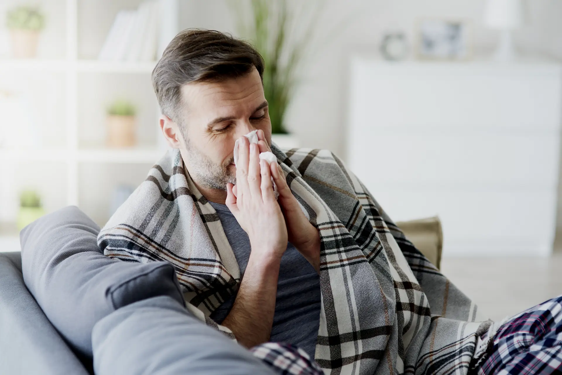 Medium shot of a person seated on a couch under a blanket blowing their nose. A sauna's infrared heat stimulates the cells in such a way that helps combat viruses. That's why sauna use is good for your immune system.